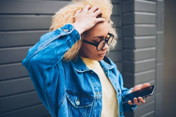 Worried young woman getting bill from service checking email on smartphone outdoors, hipster girl touching curly hairstyle solving problem with banking account sending message for customer support