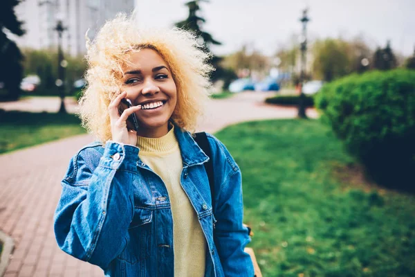 Positive woman satisfied with connection in roaming talking with friend walking on urban settings background, smiling hipster girl happy about good news getting during phone conversation outdoors