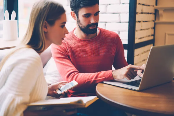 Bearded hipster guy installing on girlfriend\'s laptop new antivirus system pointing on settings while sitting in cafe, best friends paying for purchases in web store using online banking services