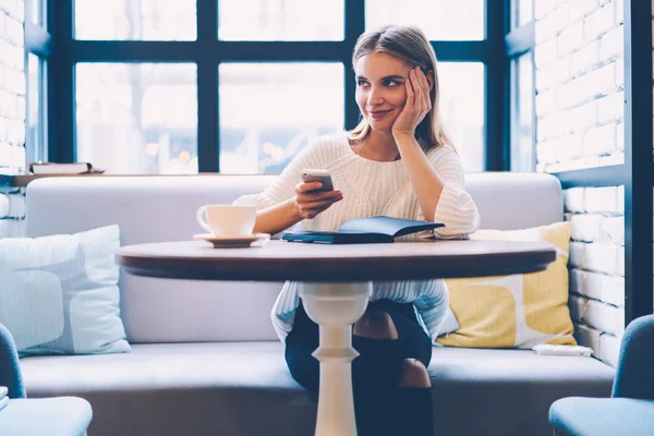 Dreamy young student resting in coffee shop during break waiting for feedback from friend holding mobile,skilled  businesswoman planning weekends while sending messages to colleague on smartphone