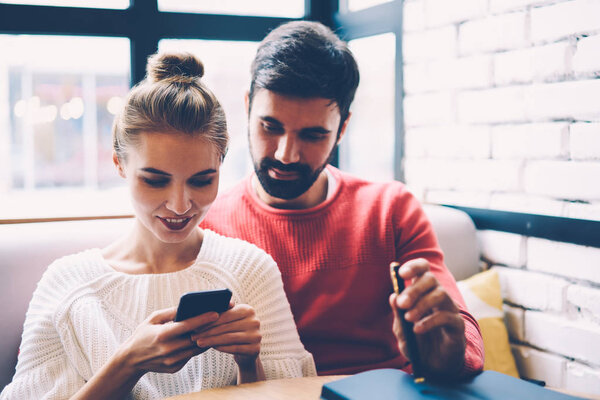 Улыбающаяся пара в любви просматривает фотографии в социальных сетях вместе отдыхающих в кафе, очаровательная подруга показывает смешное видео на мобильном с бойфрендом весело знакомства в уютном интерьере кафе
