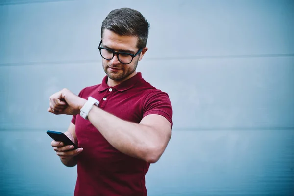 Young businessman in stylish eyeglasses with black frame holding digital smartphone in hand and looking at modern watch standing against promotional background for your advertising text message