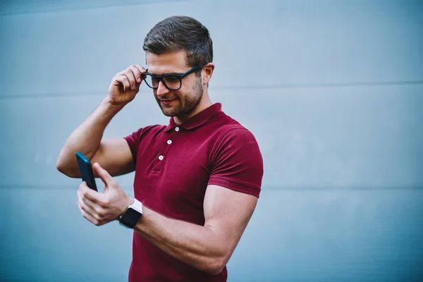 Handsome smiling young man in stylish optical spectacles reading sms message with good news on modern smartphone standing against promotional background for your advertising text message