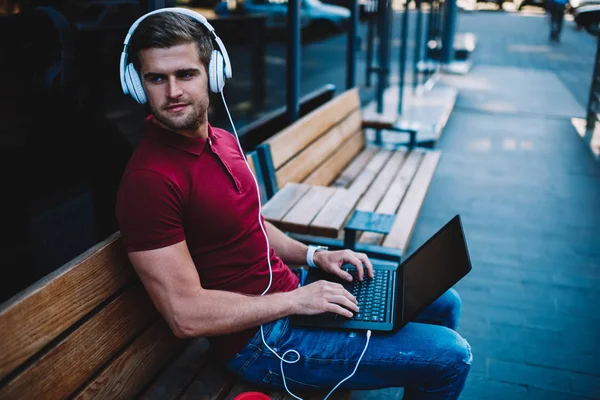 Positive handsome man resting on wooden bench with digital laptop computer and moder headphones on head.Male meloman listening favourite playlist via earphones connecting to computer outdoors