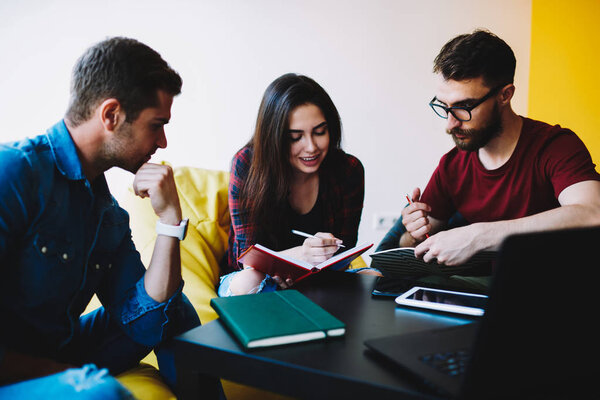 Team of skilled students collaborating on common training project in stylish coworking.Young woman with notepad in hands together with two hipster guys thinking on creative ideas sitting on poufs