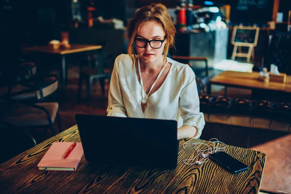 Thoughtful female copywriter concentrated on task earning money online using modern laptop, thinking businesswoman making research on web pages solving problems doing remote job in cafe interior