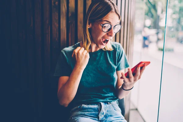 Surprised and amazed female person in cool spectacles emotionally reacting to live broadcast watching via modern cellphone indoors.Shocked female celebrating win with discounts for shopping