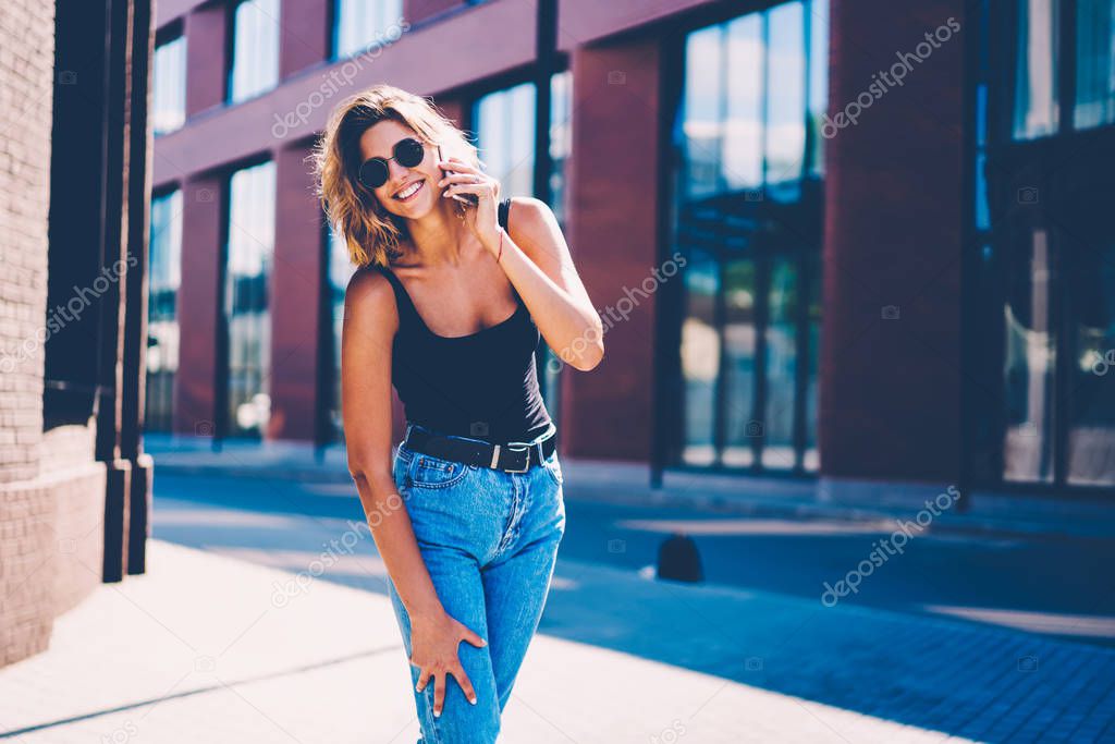 Portrait of cheerful young female in casual apparel having fun outdoors talking on mobile with friend, happy hipster girl in sunglasses excited with making calls using cheap tariffs in roaming