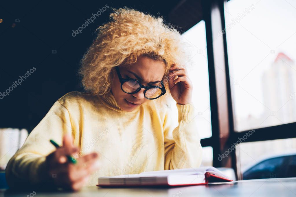 Thoughtful hipster girl in eyewear pondering on idea while reading information from notepad,contemplative young woman with curly hair analyzing details creating schedule and plan for work on free time