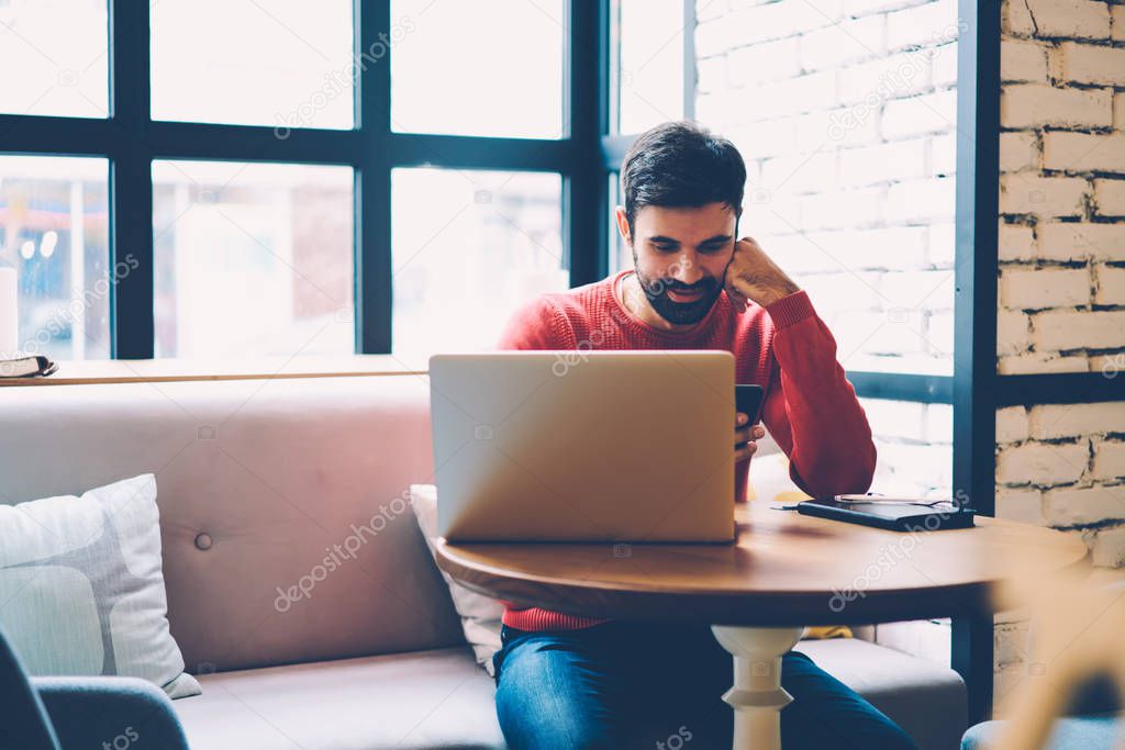 Smiling bearded guy receiving message with good news from girlfriend while doing remote job in cafe interior, skilled copywriter updating software on laptop while chatting in social networks