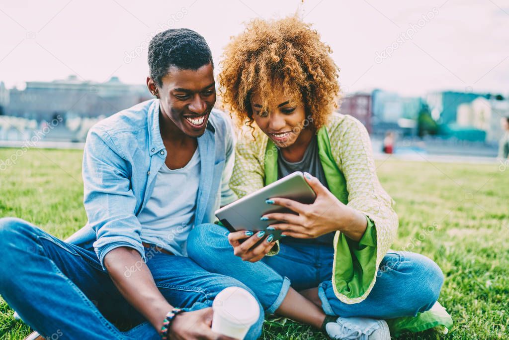 Cheerful afro american best friends watching movie online satisfied with high speed internet connection outdoors,happy couple in love laughing on funny videos on tablet sitting on lawn together