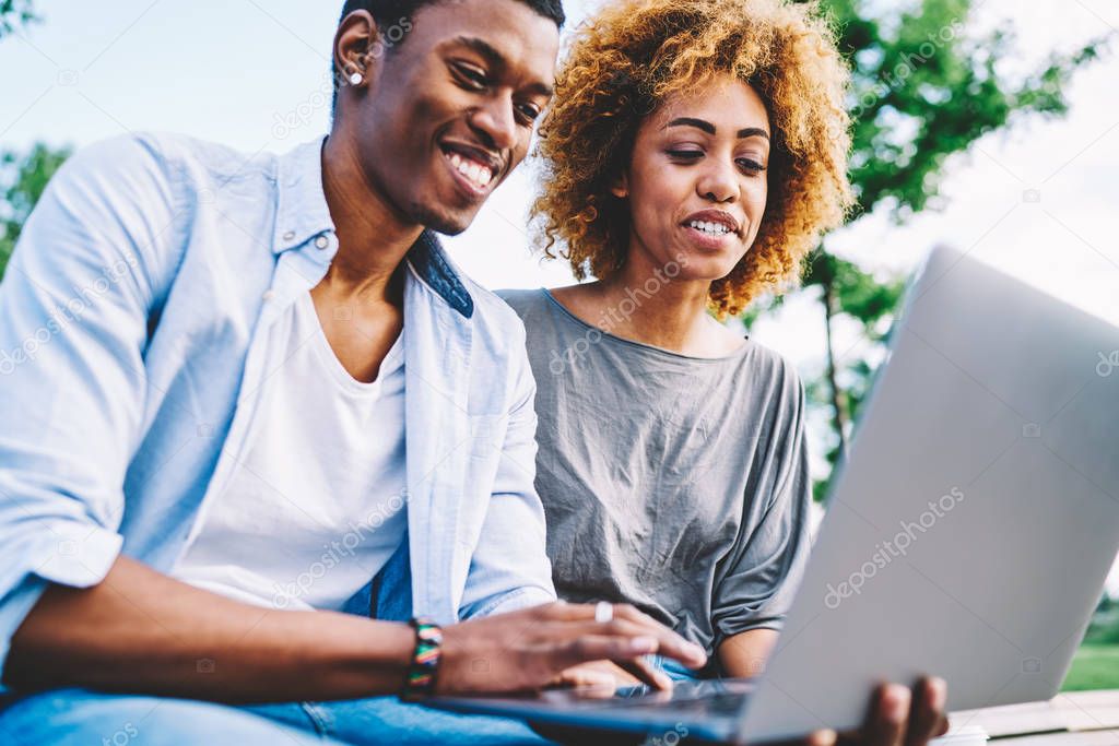 Smiling afro american friends making pictures using online camera on laptop making funny faces for share in networks, happy dark skinned hipsters enjoying resting together dating in park watching movie