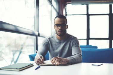 Portre konsantre erkek girişimci başarılı stratejileri notepa içinde yazma hakkında gözlük düşüncesinde afro Amerikalı işadamı modern iç kafede oturan planları çalışmak için notlar