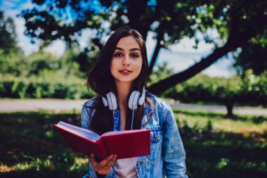 Half length portrait of charming young woman with brunette hair holding literature book in hands.Beautiful female with modern headphones on neck looking at camera while reading bestseller outdoors clipart