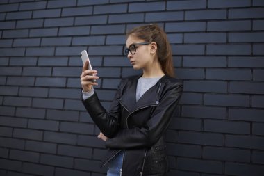 Trendy giyim ve gözlük promosyon arka plan üzerinde zaman açık havada duran harcama 4 g internet bağlantısı kullanma sosyal ağlardan video izlerken smartphone tutan genç hippi kız giymiş