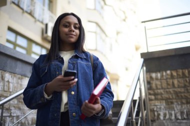 Portre çekici esmer öğrencinin Kot ceket kitapla sokak kontrol e-posta-mobil, smartphone navigasyon kullanarak toplantı için gezinme afro Amerikan hippi kız yürürken