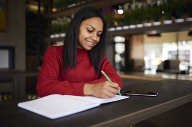 Çekici afro Amerikan kadın girişimci kahve üzerinde otururken artan gelirleri kırmak için proje planlama ve startu için yaratıcı fikirler belirterek neşeli Yöneticisi günlüğünde listelemek yapma