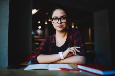 Yarım uzunluk portre esmer çekici öğrenci şık Eyewear kafede tahta masada otururken kameraya bakıyor. Boş vakit kapalı çekici genç hippi kız