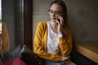 Güzel genç moda gözlük modern smartphone konuşuyor. Şık coworking uzayda oturan telefonu iletişim gözlük sarı saçlı sevimli genç kadın