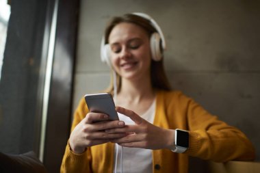 Modern cep telefonu hangi gülümseyen hipster adam elinde tutan ve kapalı alan coworking oturan şık kulaklıklar ile en sevdiğiniz müzik dinleme sırasında sosyal ağlarda arkadaşlarınızla çevrimiçi sohbet tarih seçici odak