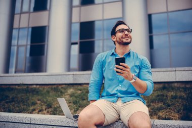 Hafta sonları planlama ve Smartphone multimedya dosya indirme Üniversitesi kampüsünde dinlenme mobil, neşeli öğrenci üzerinde uygulama üzerinden online bilet rezervasyonu trendy gözlük mutlu hipster adam