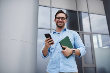 Trendy Eyewear açık havada dizüstü bilgisayar, kamera iletileri göndermeyi fon üzerinde seyir yakışıklı hipster adam ayakta iken Smartphone multimedya dosya indirme başarılı işadamı portresi