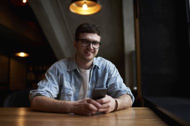 Kahve Dükkanı iç otururken modern telefonda uygulama kullanarak trendy optik gözlükler hipster adam portresi. Adam smartphone cep telefonu ile sosyal ağ Haberler okuma kot gömlek giymiş