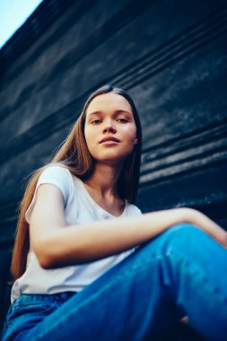 Kadın, doğal güzellik makyaj moda giyim fotoğraf için poz olarak çekici kamera duvar arka plan açık havada oturup raslantı giyimli çekici hipster kız portresi