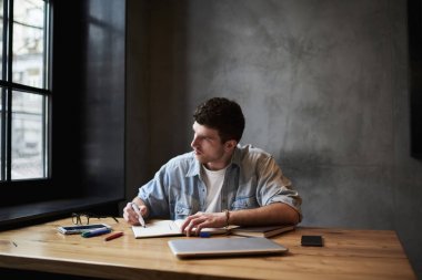 Genç yetenekli mimar marker elinde tutan ve Not Defteri'nde studio iç ahşap masaüstü çalışma işlemi sırasında oturup pencereden dışarı bakarken planlama inşaat kroki çizim