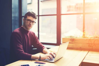 Profesyonel erkek portresi bu kameraya bakarken güneş ofis iç pencere yanında oturan modern dizüstü cihaz üzerinde yeni programlar oluşturma konusunda çalışma gözlük geliştirici