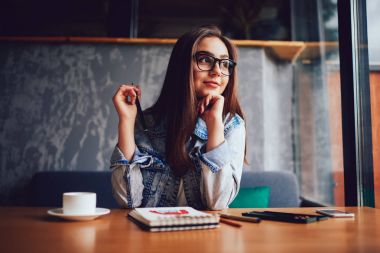 Rüya gibi çekici öğrenci şık optik gözlükler fincan kahve ile ahşap masa kafeteryasında oturan ve Not Defteri'nde eskiz çizmek için yeni yaratıcı tasarımcının fikir üzerinde düşünüyorum