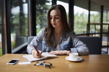 Akıllı yetenekli öğrenci ödev alan coworking içinde kahve ile masada oturan not defteri olarak aşağı yazma esmer saçlı. Not defterinde metin bilgileri belirterek dalgın genç kadın
