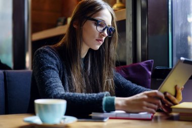 Akıllı kız öğrencinin not defterinde kahvehane iç ile oturan dijital dokunmatik yüzey üzerinde bilgi aramak. Şık gözlük kafede modern tablet metin okuma akıllı genç kadın