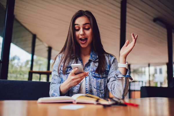 Эмоциональная хипстерша получила шокированные новости на современном смартфоне. Удивлена, что женщина читает смс на цифровом телефоне во время чтения интересной книги в коворкинге
