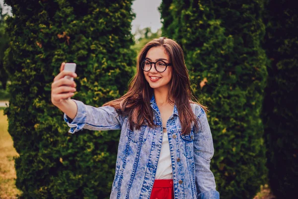 Şirin esmer hippi kız modern telefon açık havada duran üzerinde selfie yapma. Fotoğrafları yayımlama sosyal ağlar için Smartphone yapma trendy denim ceket giymiş neşeli genç kadın