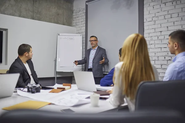 有经验的男性营销专家在眼镜示意 同时解释给梦想团队成员工作过程中协作 在办公室互相沟通的设计师组 — 图库照片