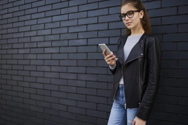 Half length portrait of teen hipster girl in eyeglasses dressed in stylish outfit blogging in social networks sharing multimedia information using 4g internet connection standing outdoors on street