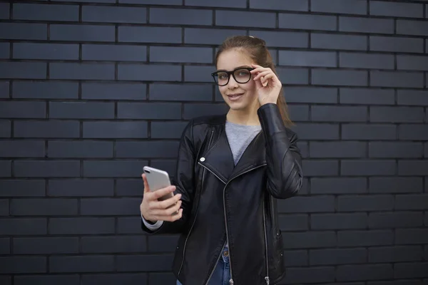 Portrait of smiling female blogger dressed in casual trendy wear holding smartphone surfing internet and chatting via 4G connection, young woman in eyeglasses looking at camera standing on copy space