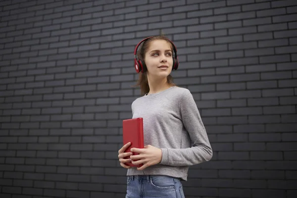 Pensive casually dressed female student strolling with notebook in headphones enjoying radio broadcasting, hipster girl in sweatshirt listening to music standing on copy space area for advertising