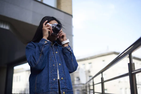 Skilled afro american female photographer in trendy denim jacket taking photo on vintage camera working outdoors, young dark-skinned hipster girl making photo of urban settings spending time on hobb