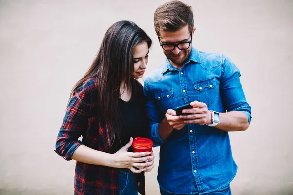 Positive stylish hipster guys watching funny photos on smartphone device.Young woman with coffee in hand and smiling boyfriend publishing pictures in social networks on telephone via free 4G internet