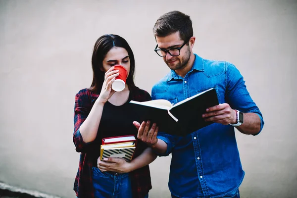 Skilled students reading training book during coffee break and preparing for next exam standing against promotional background.Hipster guys learning text information from textbook in free time