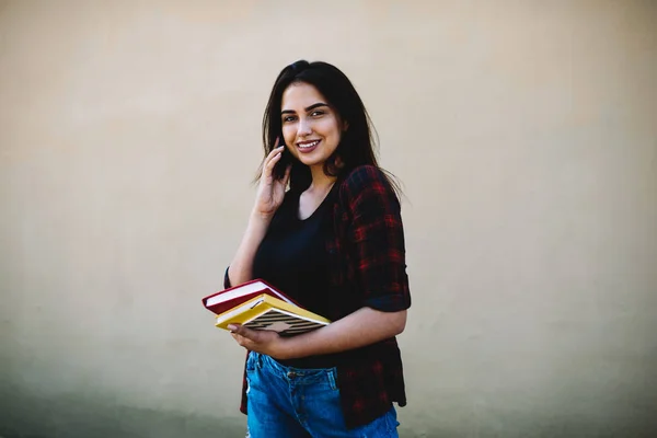 Half length portrait of prosperous talented student looking at camera while communicating with friends via cellular.Pretty hipster girl using technology during standing against wall with copy space