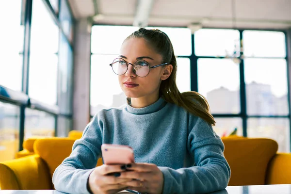 Uzakta pencere bloglama ağlar yakın oturan ve mobil multimedya dosya indirme arayan çekici genç öğrenci. Dalgın işkadını telefon kafede oturmuş bekliyor