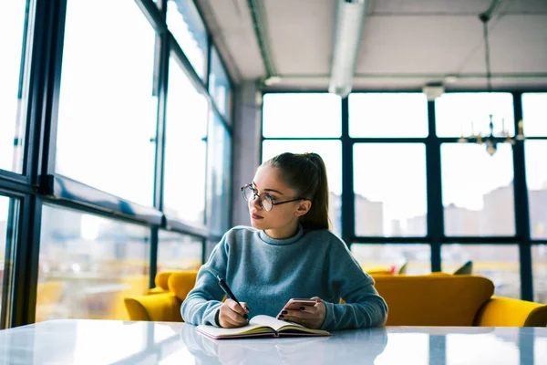 Modern Kütüphane bilgileri öğrenme pencere bakarak Eyewear dalgın genç öğrenci ödev görev üzerinde yoğunlaştı. Çekici kadın yazar iş düşünmeye smartphone üzerinde uygulama güncelleştiriliyor