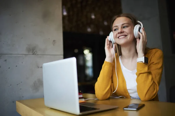 Portrait of cute blonde student enjoying while listening favorite songs via player on laptop sitting in cafe.Positive hipster girl fond of music feeling excited with perfect sound in headphones
