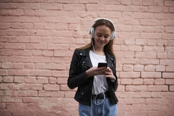 Positive blogger publishing new story in social networks on smartphone while enjoying favourite music via headphones.Hipster girl listening player on mobile phone standing on promotional background