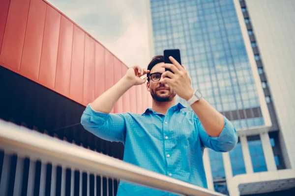 英俊的时髦的家伙在时髦的眼镜摆在自拍站在背景与当代城市建筑 年轻的游客在手机上做博客在社交网络上的图片 — 图库照片