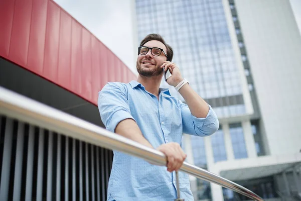 Smiling male tourist in eyeglasses satisfied with good international connection while talking on mobile with friend, handsome hipster guy enjoying phone conversation having cheap tariffs in roamin