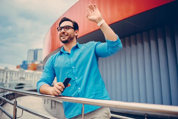 Positive young hipster guy in trendy casual outfit waving fro girlfriend waiting for date standing near modern constructed building, happy bearded man in eyewear raising hand to say hello to somebody